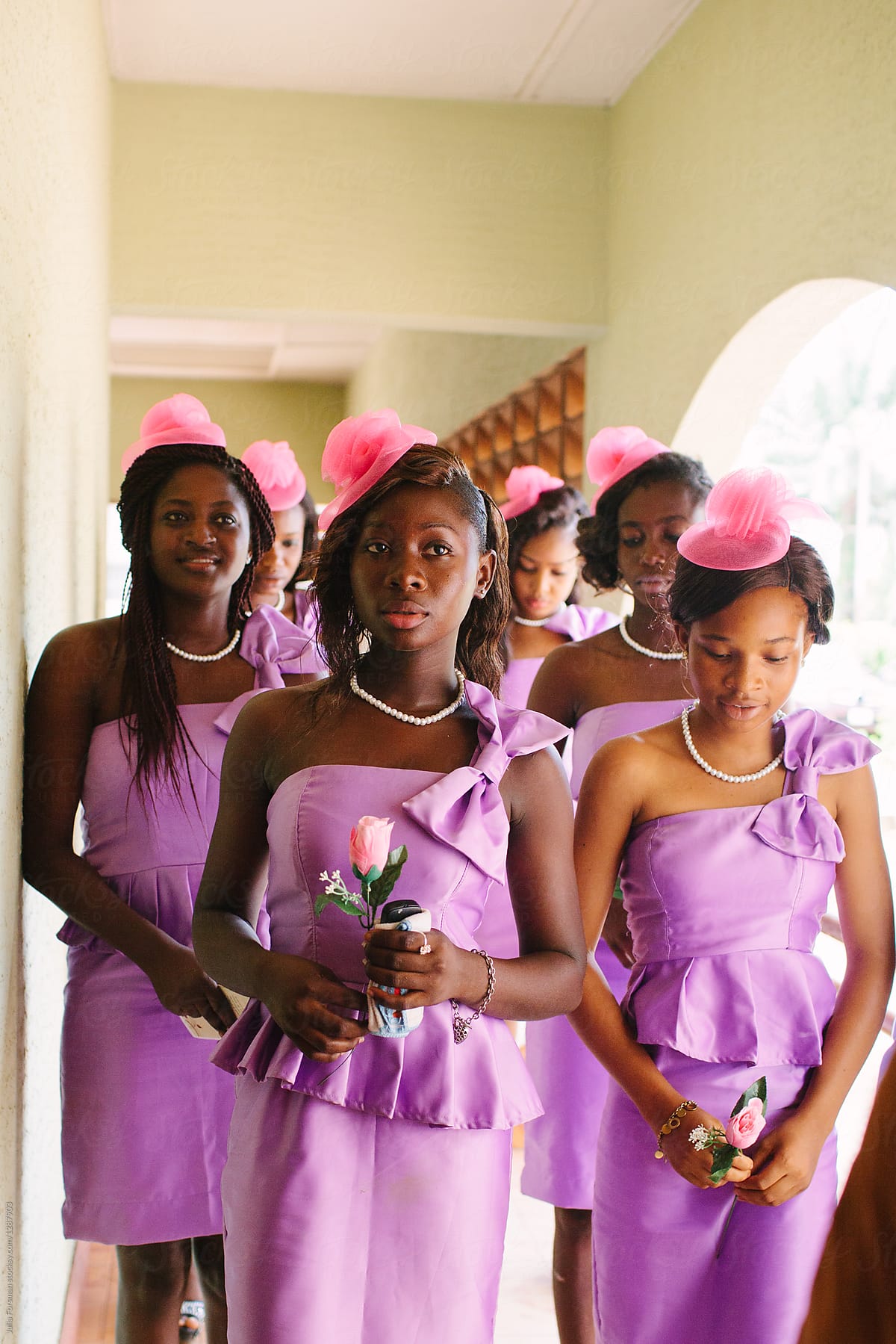 A group of six African bridesmaids wearing purple dresses and pink hats stand waiting before the wedding.