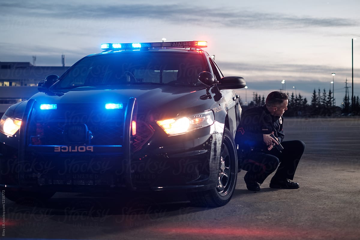 A Police Officer crouches next to a cruiser with his gun drawn