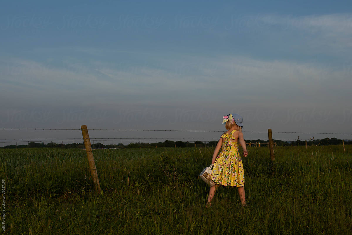 A little girl in a summer dress and sun hat takes a walk through a field.