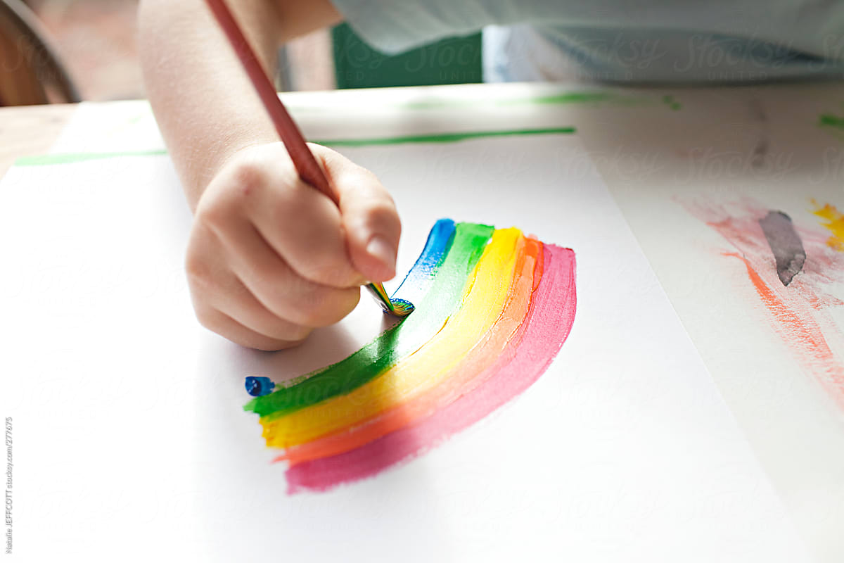 A young boy paints a rainbow at a table in the afternoon window light