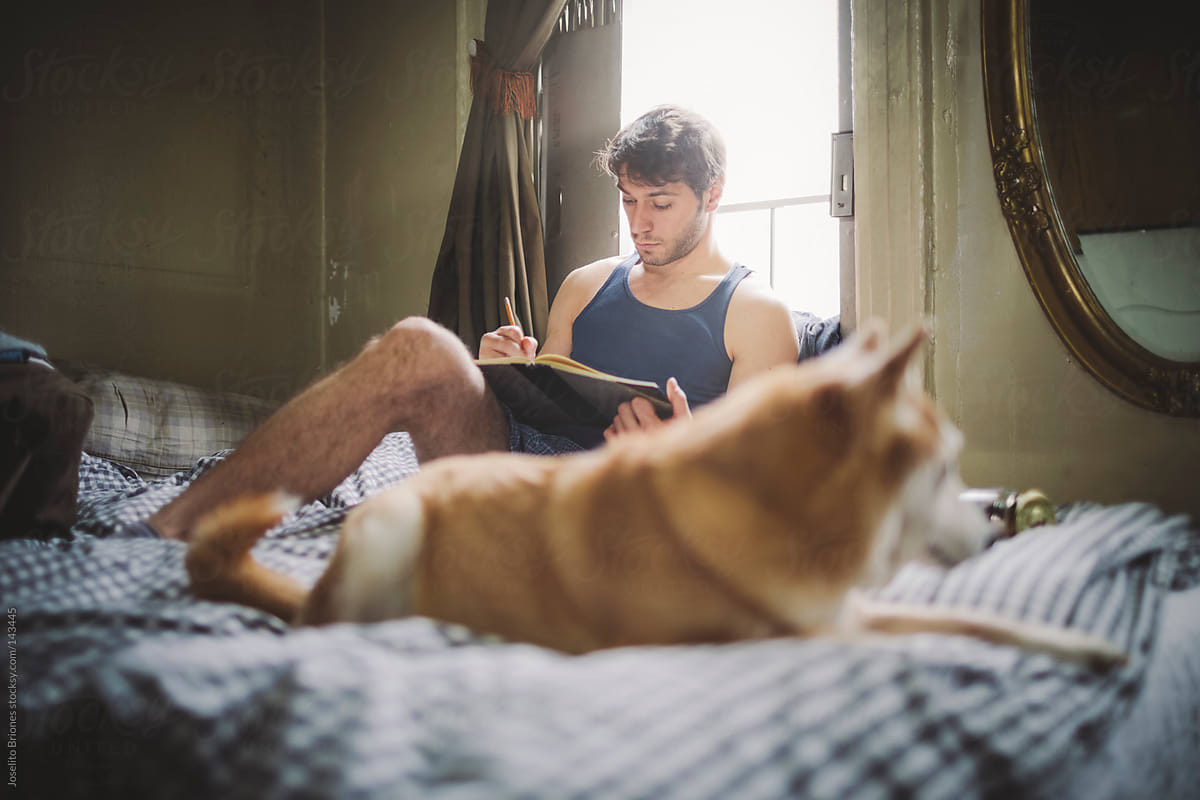 Man Writes in Notebook in Bed at Home with Pet Dog