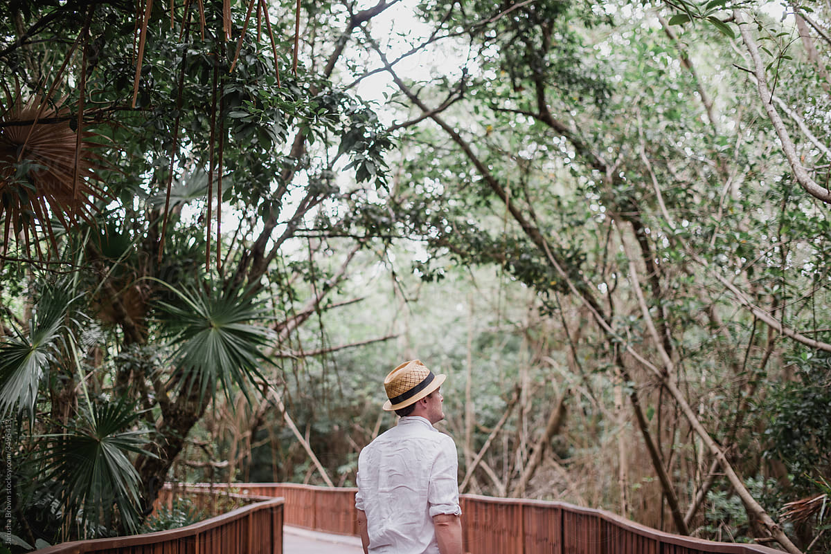 Man in straw fedora walks on a boardwalk in the jungle.