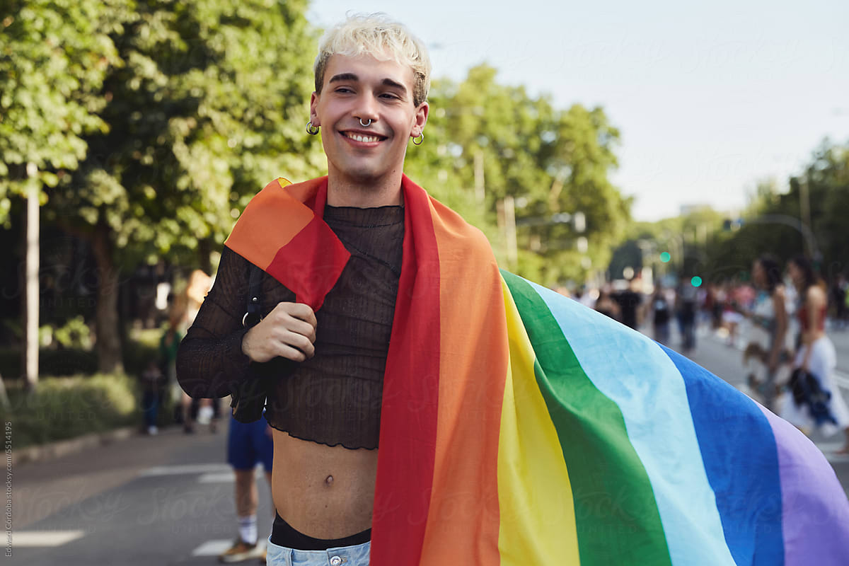 A young blonde guy proudly holding up an LGBTIQ+ flag.