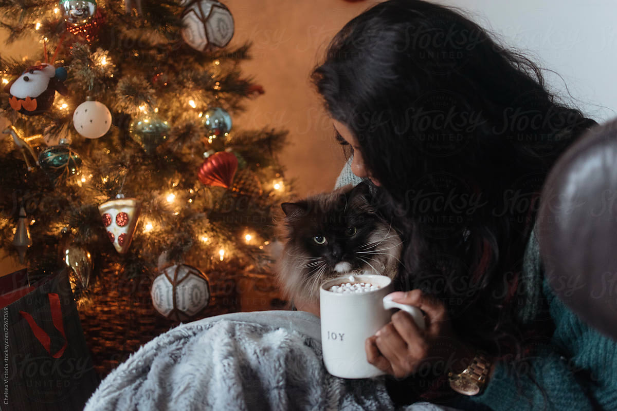 A woman sitting by a christmas tree sipping hot cocoa with her cat