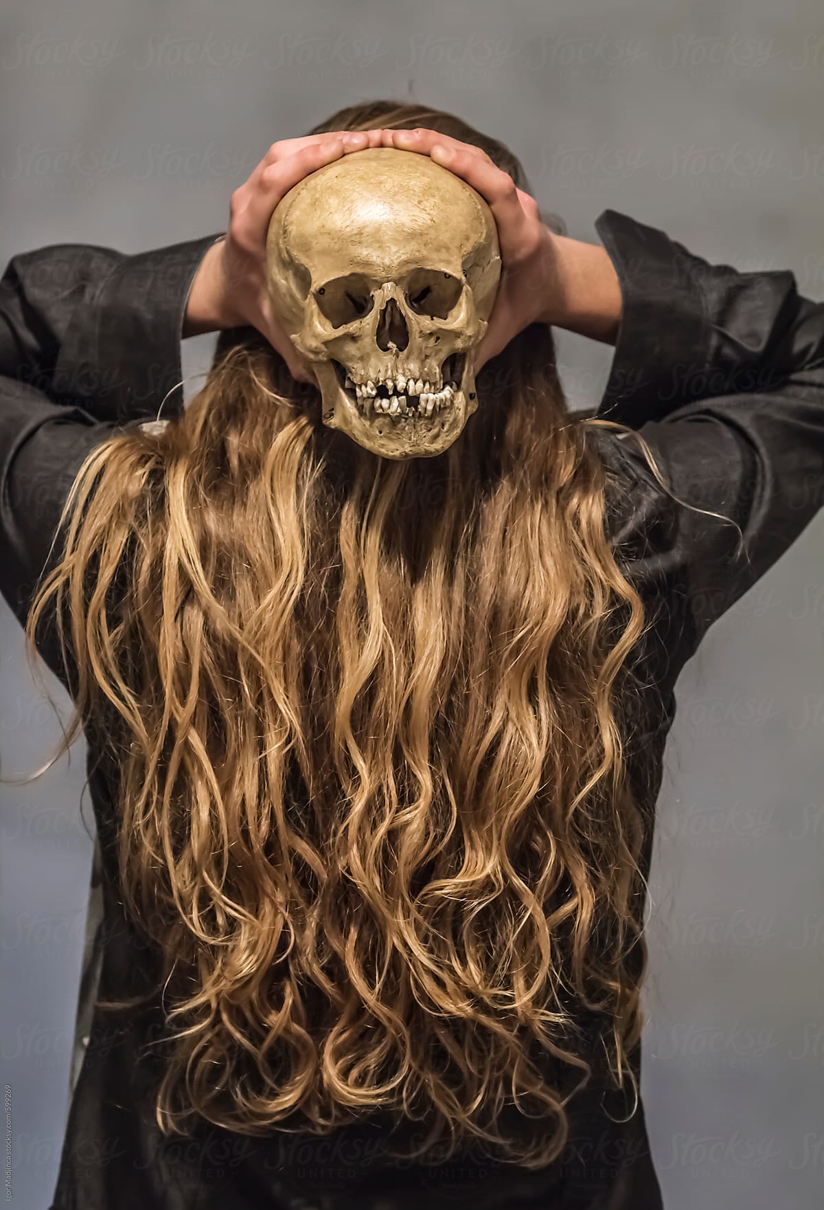 long-haired blond man from behind, holding the skull vertically
