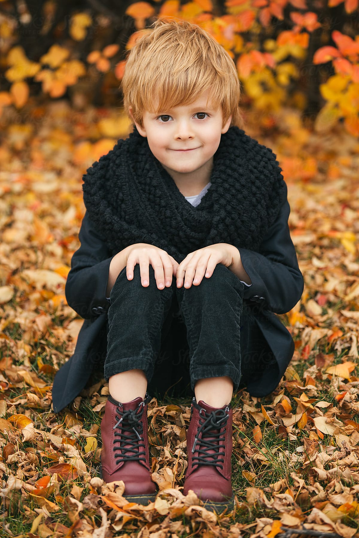 A young boy sitting on a ground covered with fall leaves smiling at camera