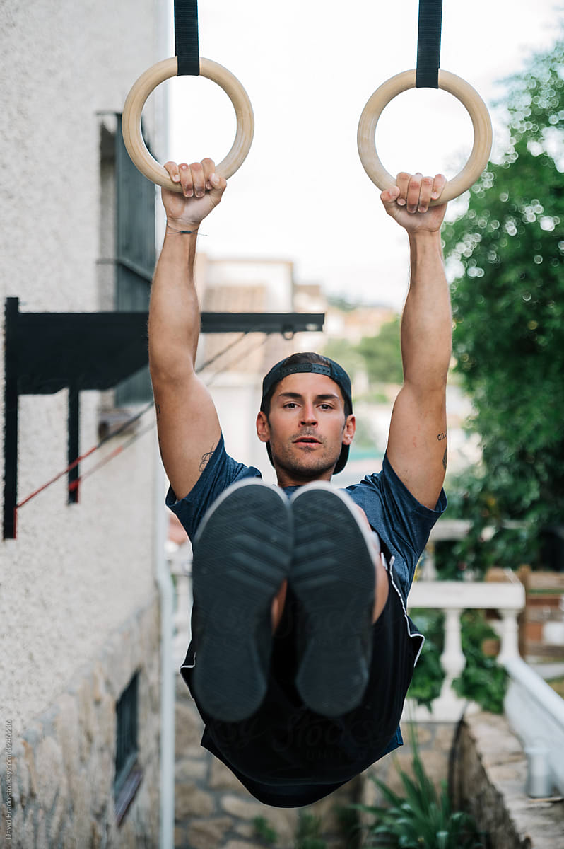 Athletic young man exercising on gymnastic rings near house