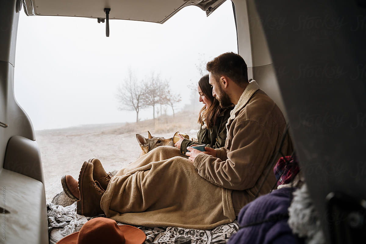 Young couple in a camper van in nature during winter with cute dog having warm coffe