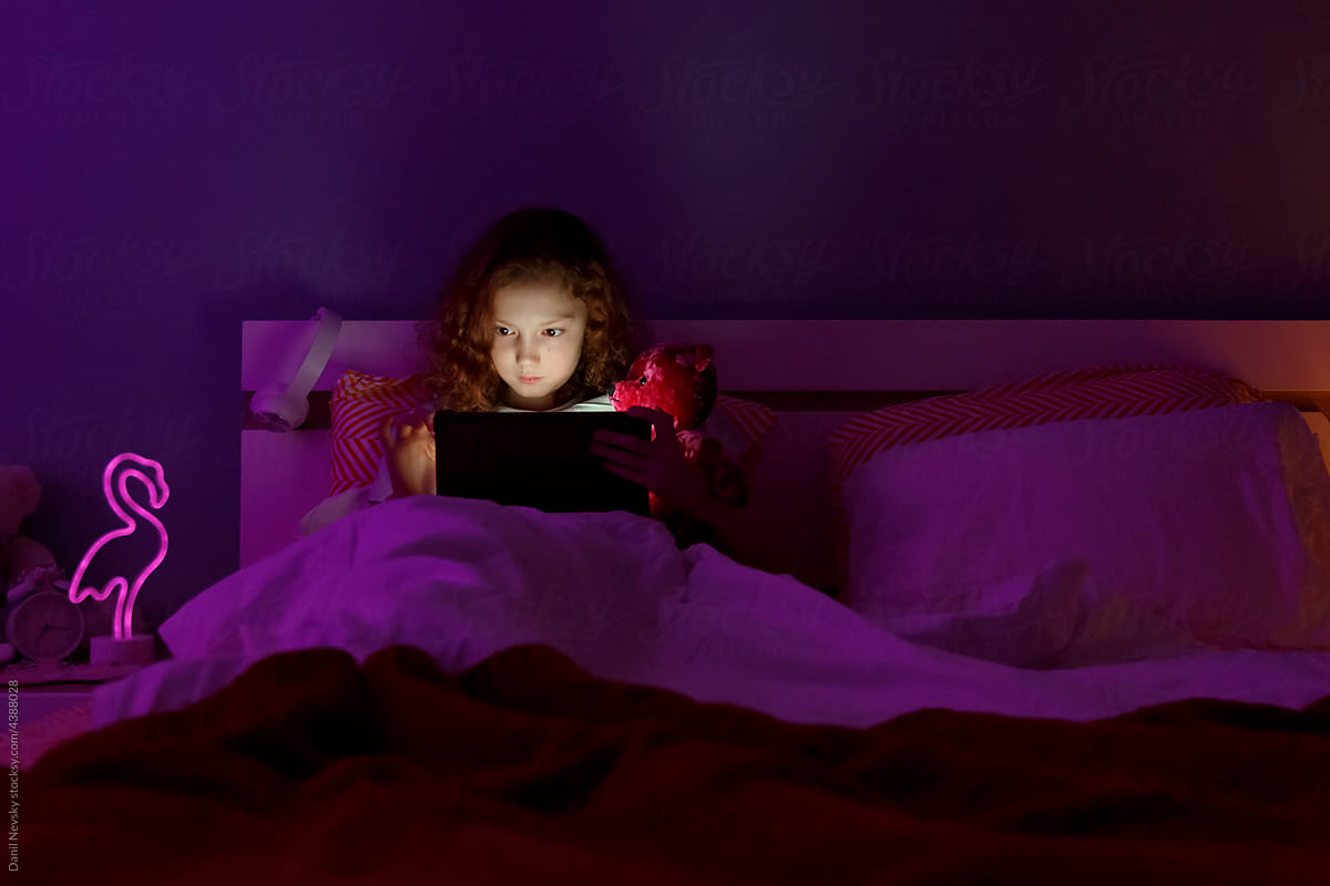 Child with teddy bear using tablet in dim bedroom