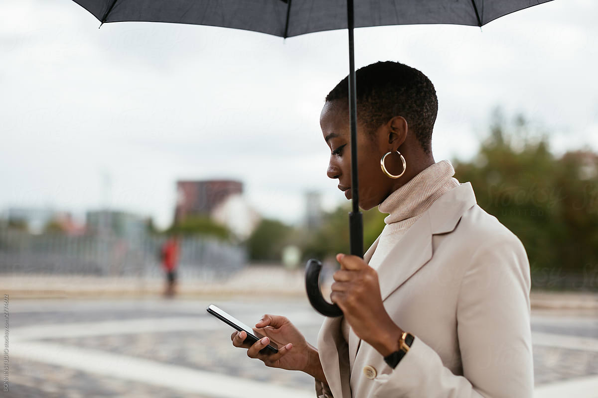 African American woman with umbrella using smartphone in park