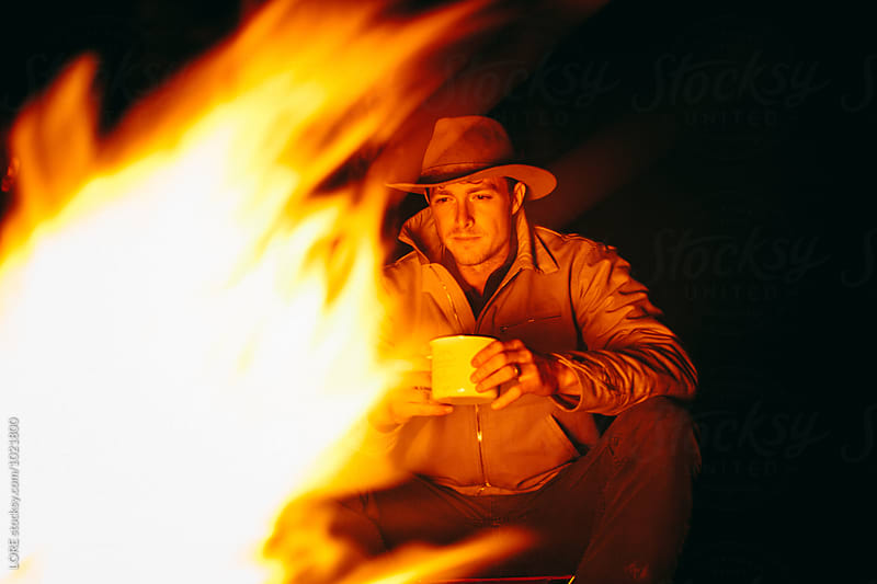 an adult male hunter sits thoughtfully by the fire at night and drinks