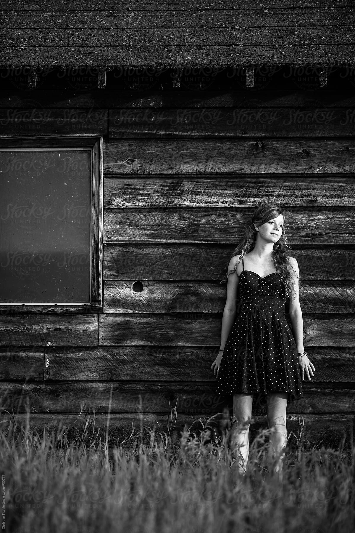 Black and white of young girl wearing a dress leaning against a rustic barn looking to the right