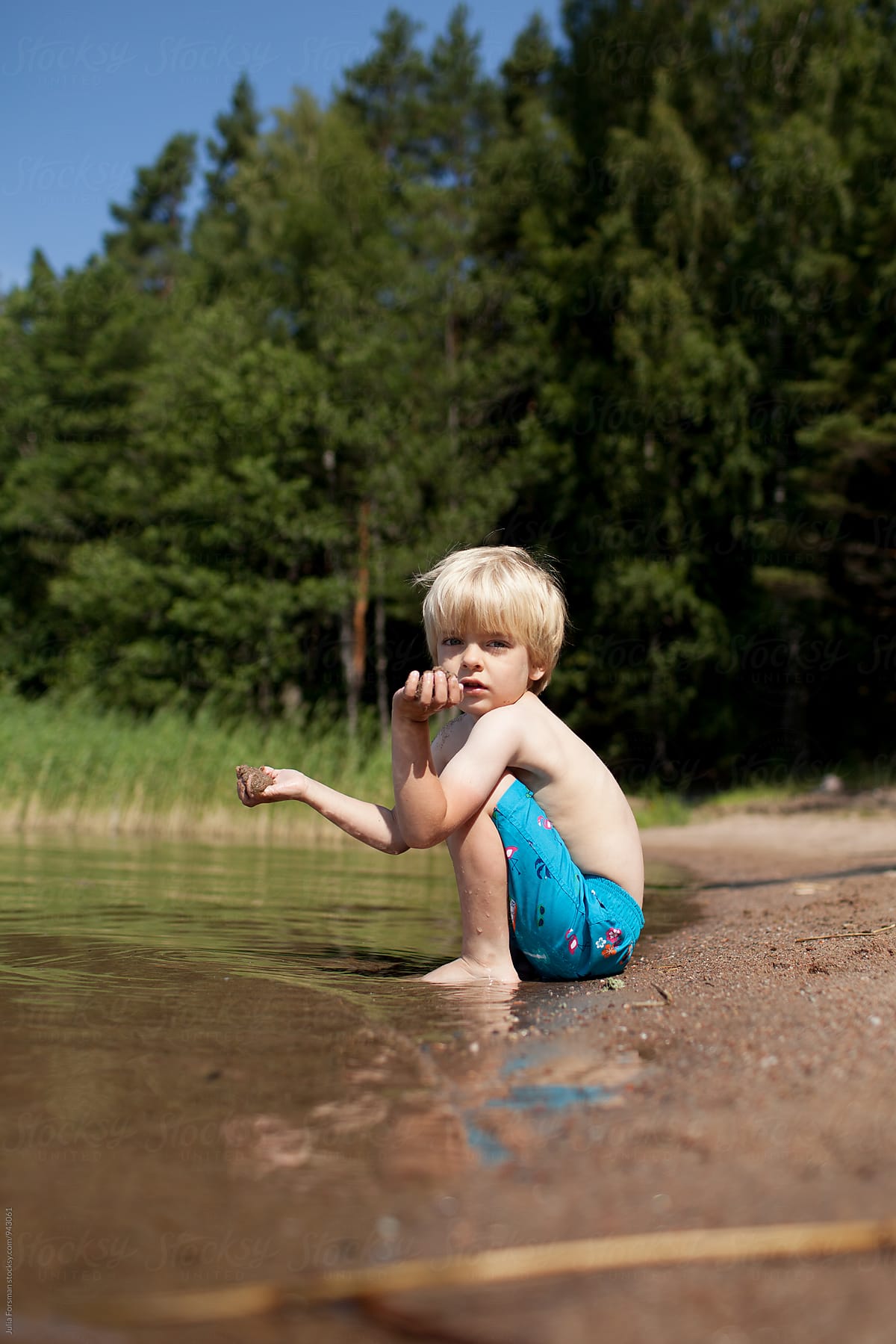 Scandinavian child sitting on a beach with forest behind him.