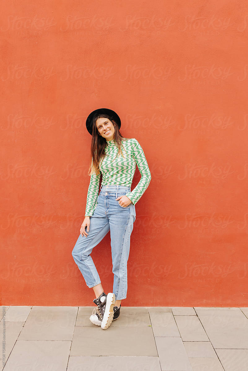 Stylish woman in hat and denim outfit standing near red wall