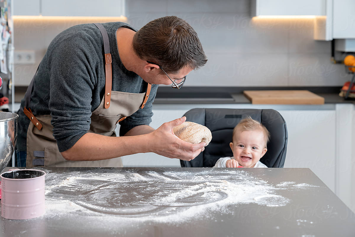 Father showing bread dough to a smiling baby in the kitchen.