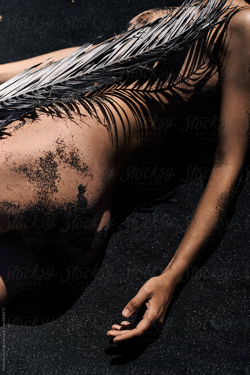 nude woman lying on the black sand and covering body with palm branch