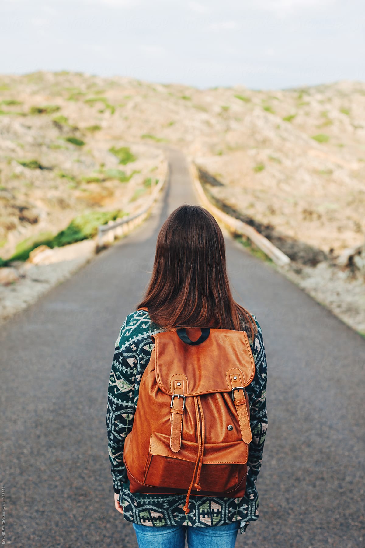 Back view of young woman with leather backpack in the middle of the road.