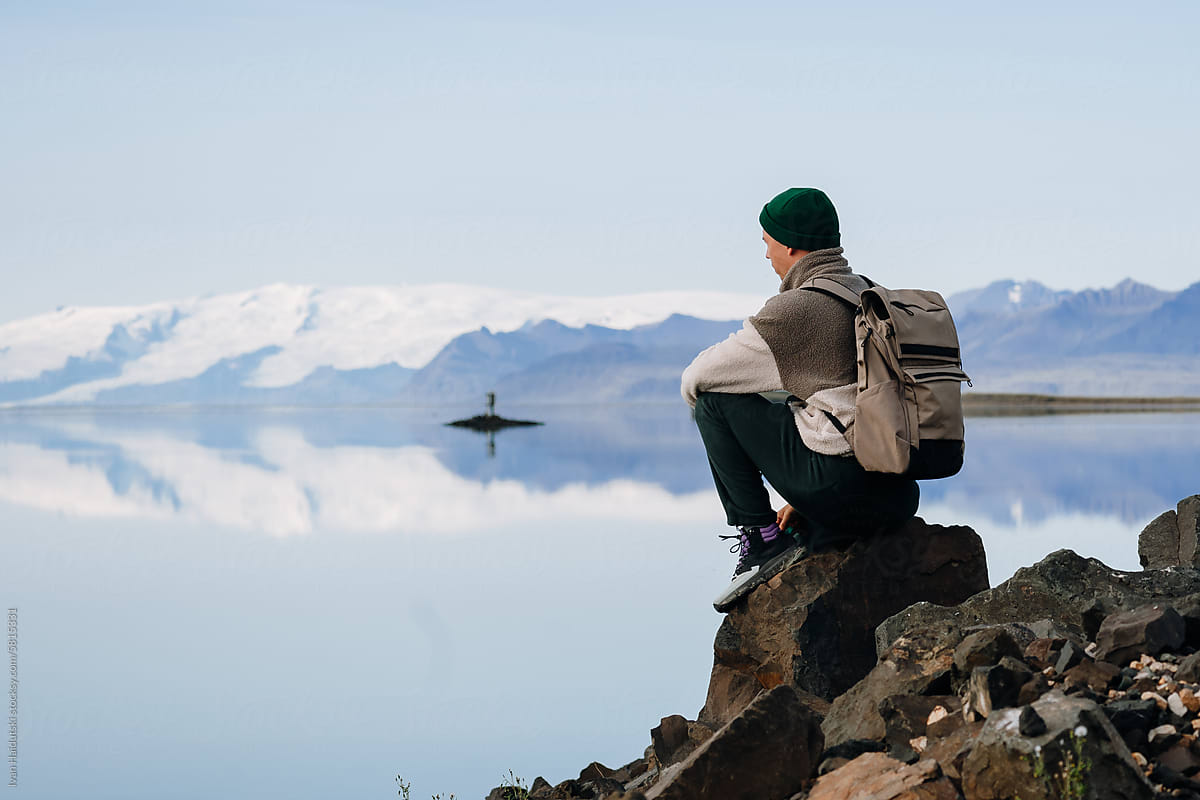 Man sitting by glacier lake shoreline. Snowy mountain water reflection