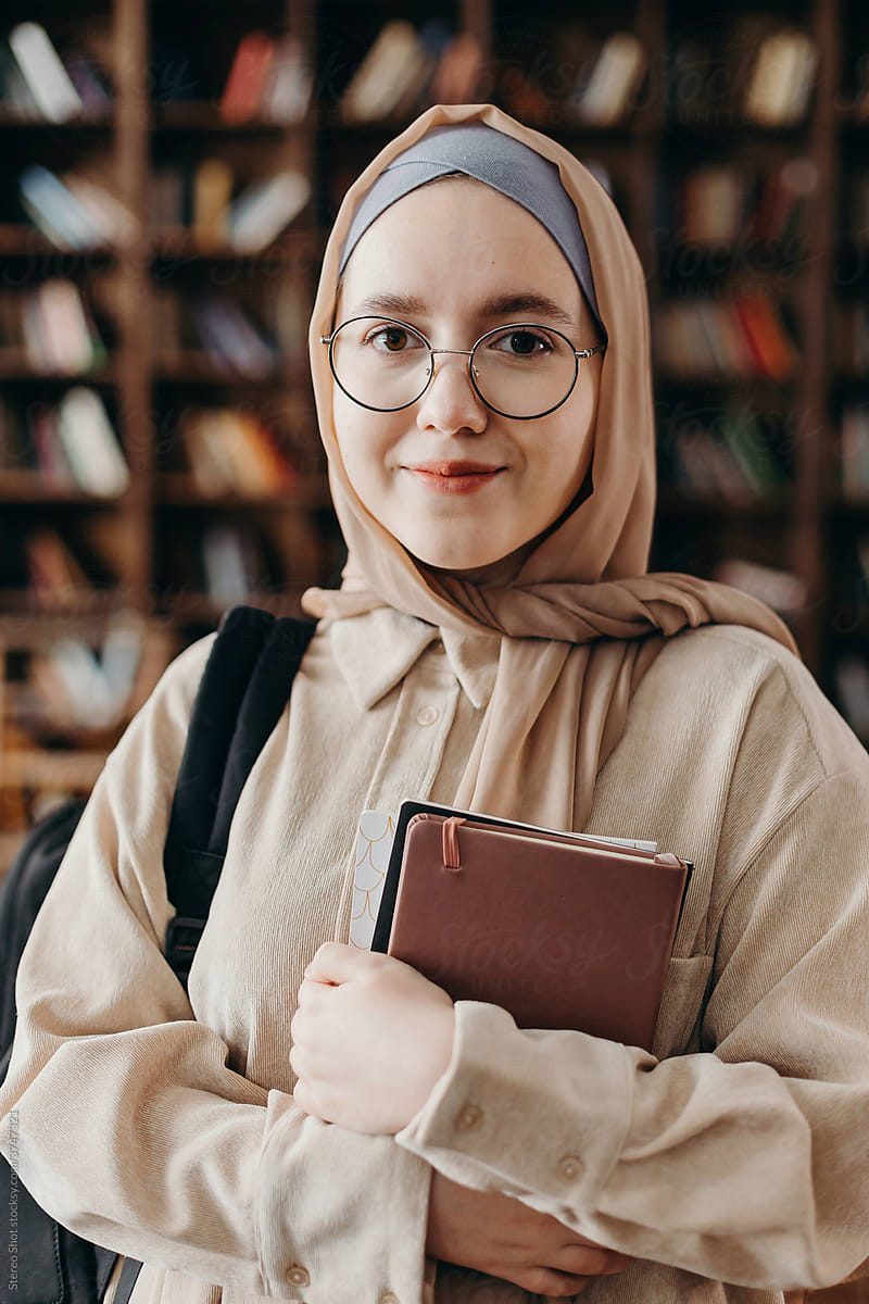 Positive Muslim teenage student standing with book in college library