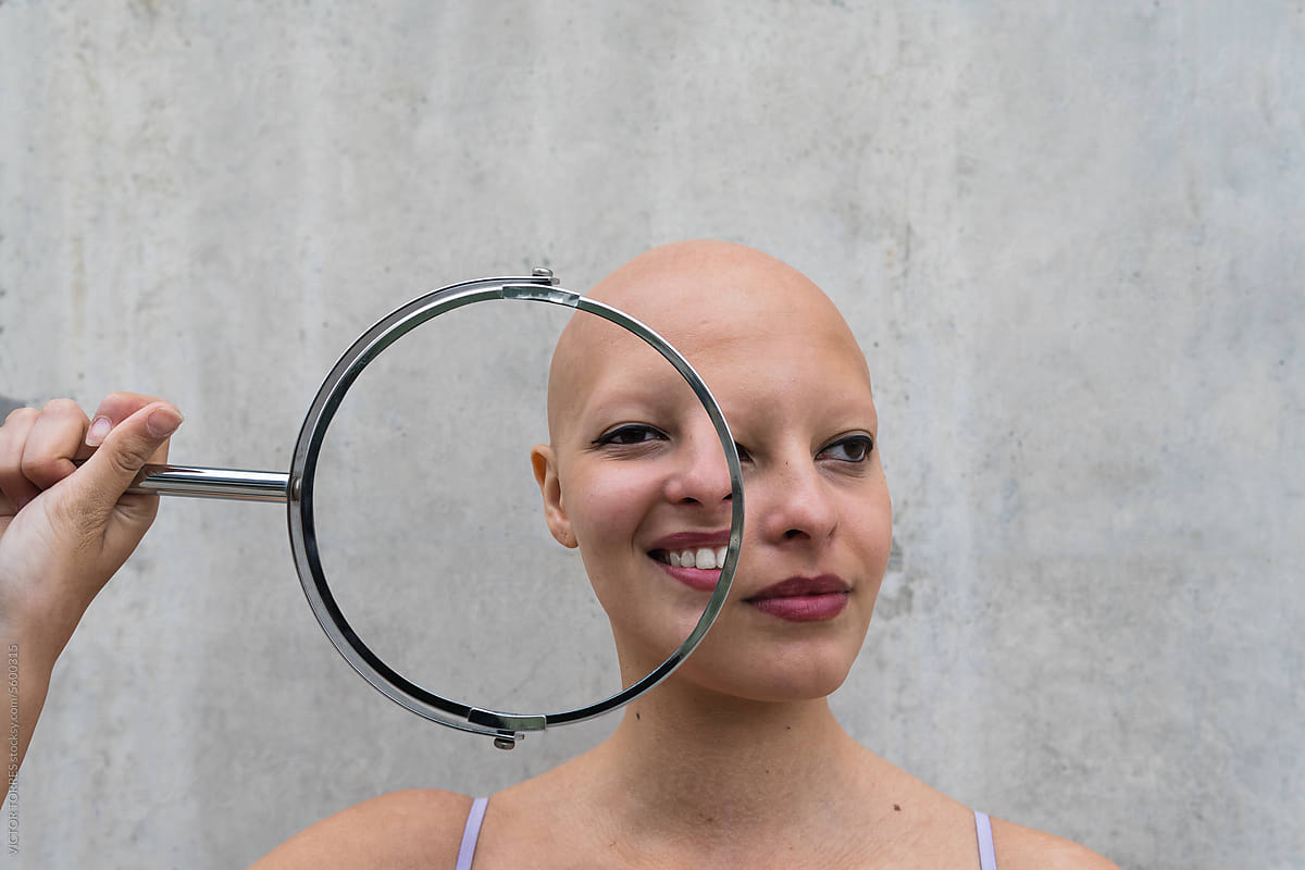 Pensive hairless woman holding mirror near face showing differen