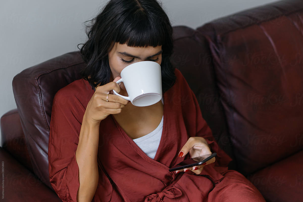 Woman sitting on a couch, drinking a coffee and browsing on a mo