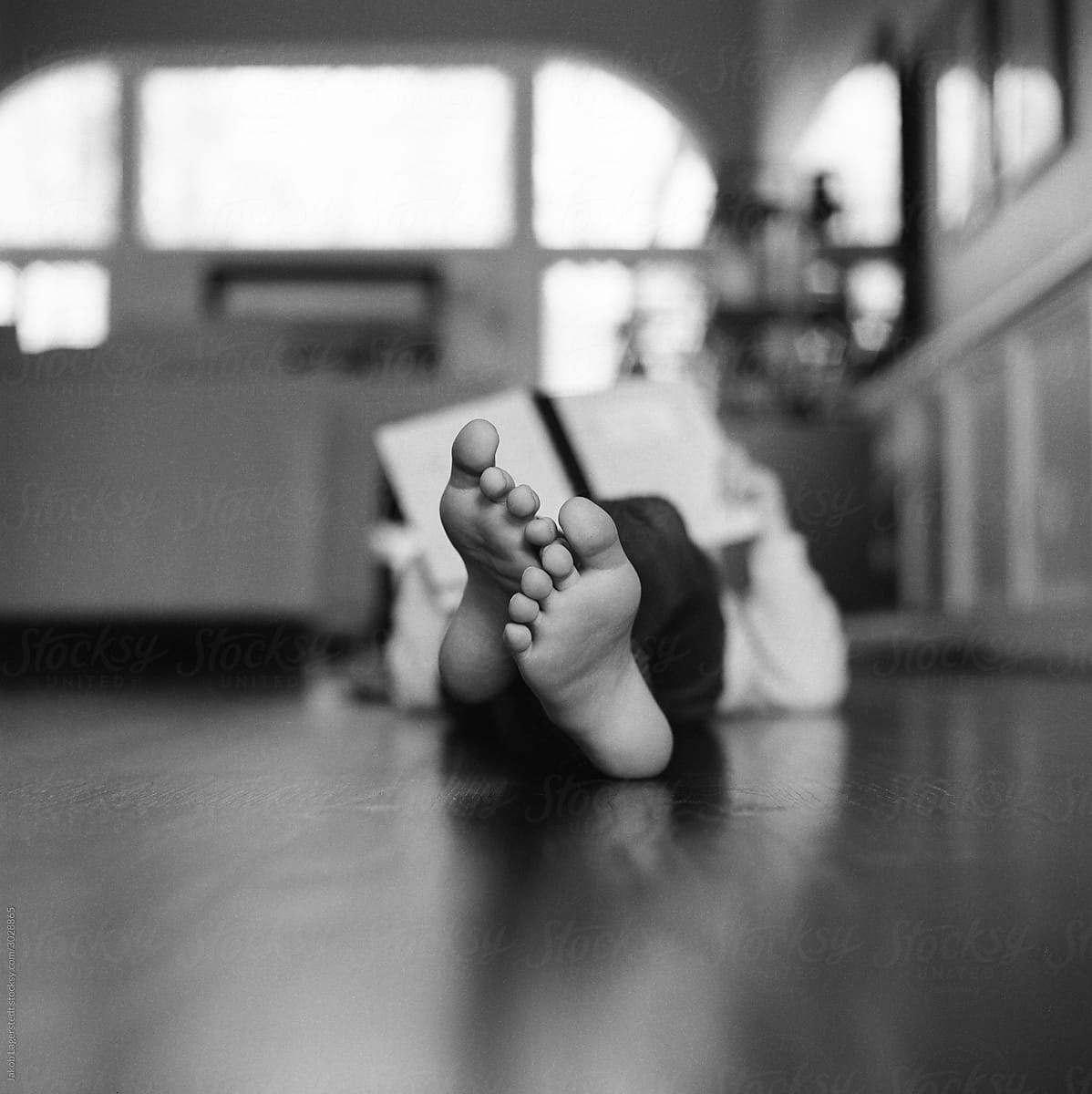 Young girl laying on hardwood floor reading a book with only her feet in focus.
