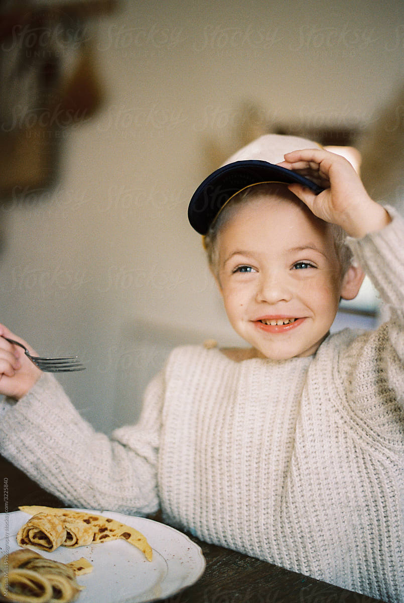 Toddler boy at the kitchen table eating pancakes and lifting his cap with one hand