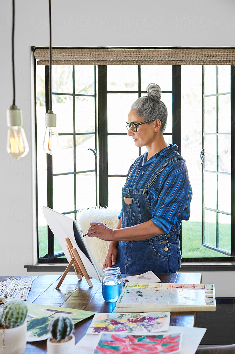 Mature woman artist with grey hair in her art studio in California painting a watercolor
