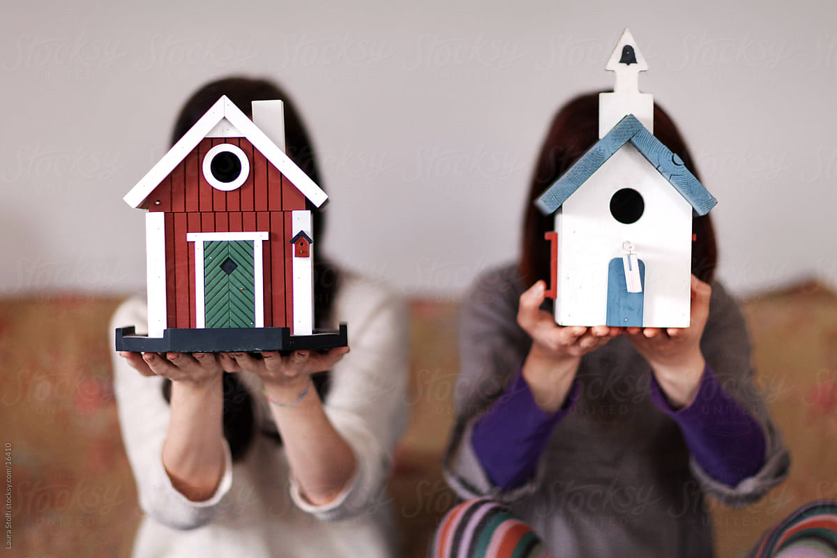 Two young women sitting on bed and hiding their faces behind barn and church shaped wooden bird houses