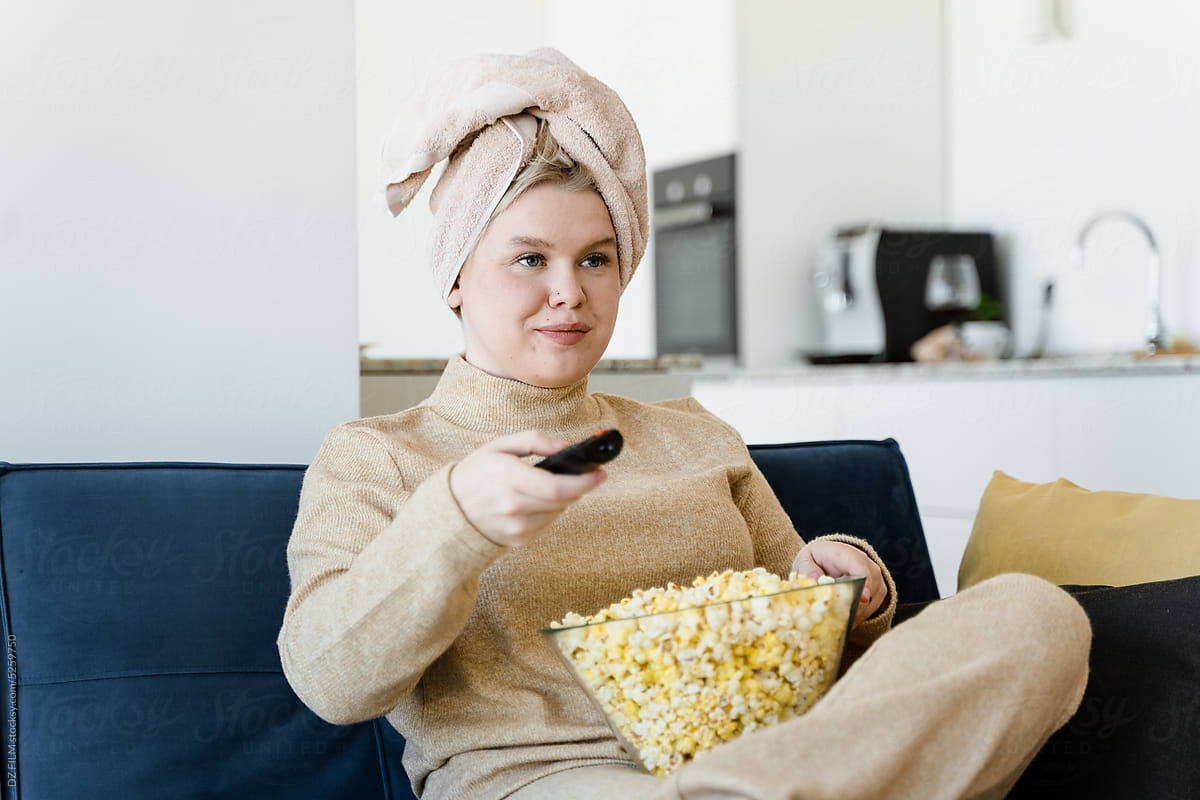 A woman with a TV control panel and popcorn sits on the couch