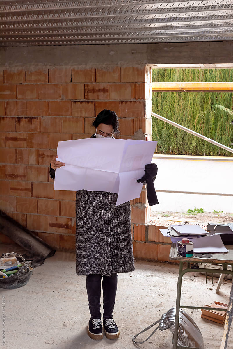 Woman in protective mask reading blueprint of new house