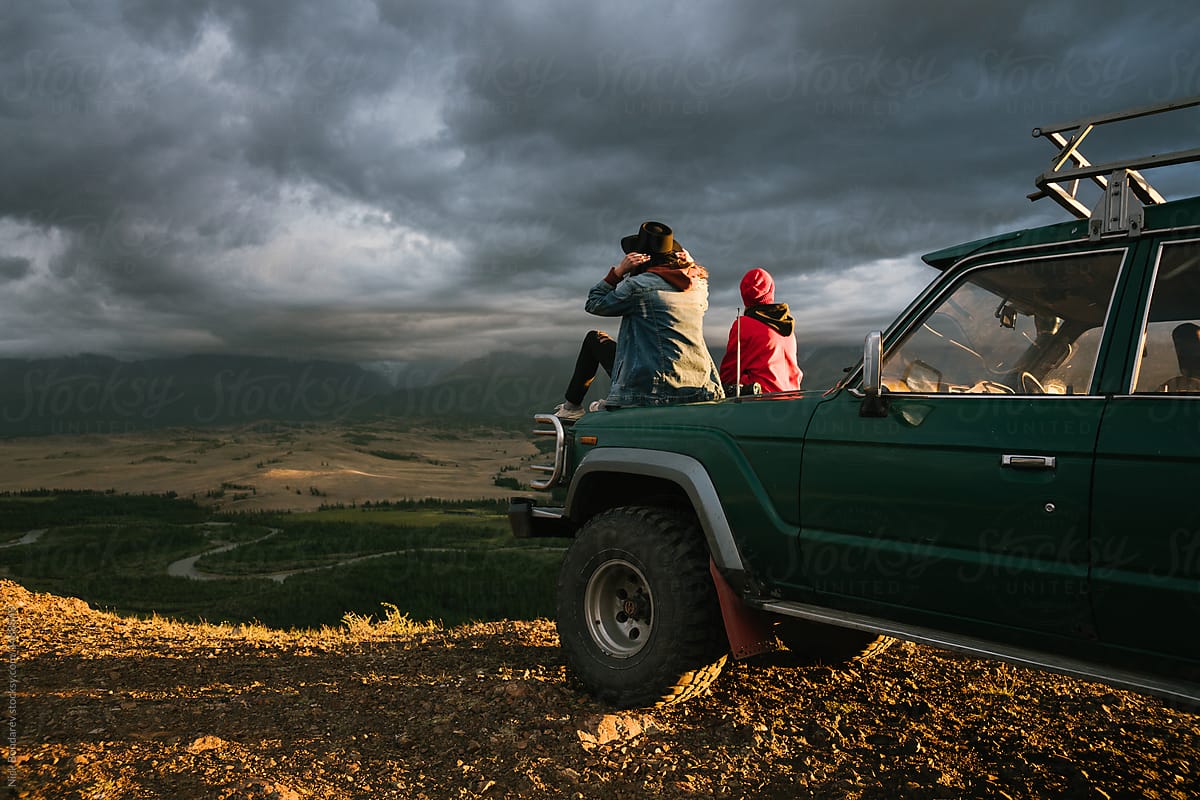 Two anonymous girls sitting on old jeep parked in the wild area surrounded by mountains