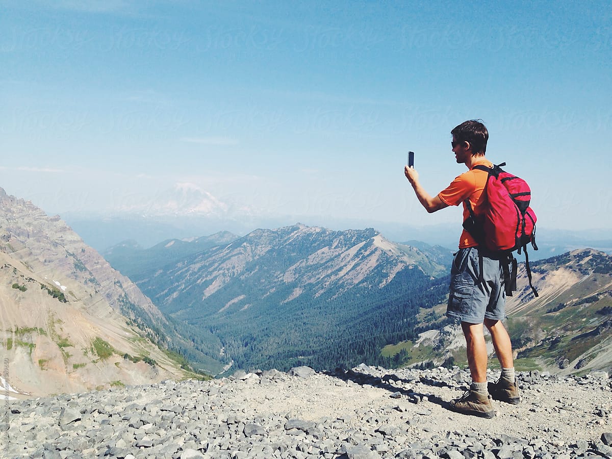 Male hiker standing on mountain summit, taking photographs with smart phone