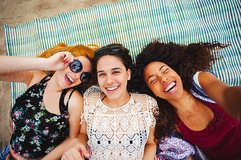 Overhead Of Smiling Female Friends Taking A Selfie Lying On The Beach 