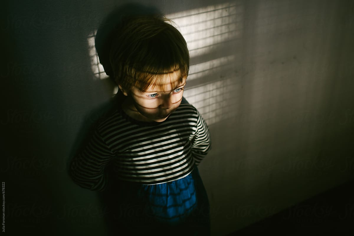 A little girl stands against a wall waiting in a patch of light containing criss cross shadows.