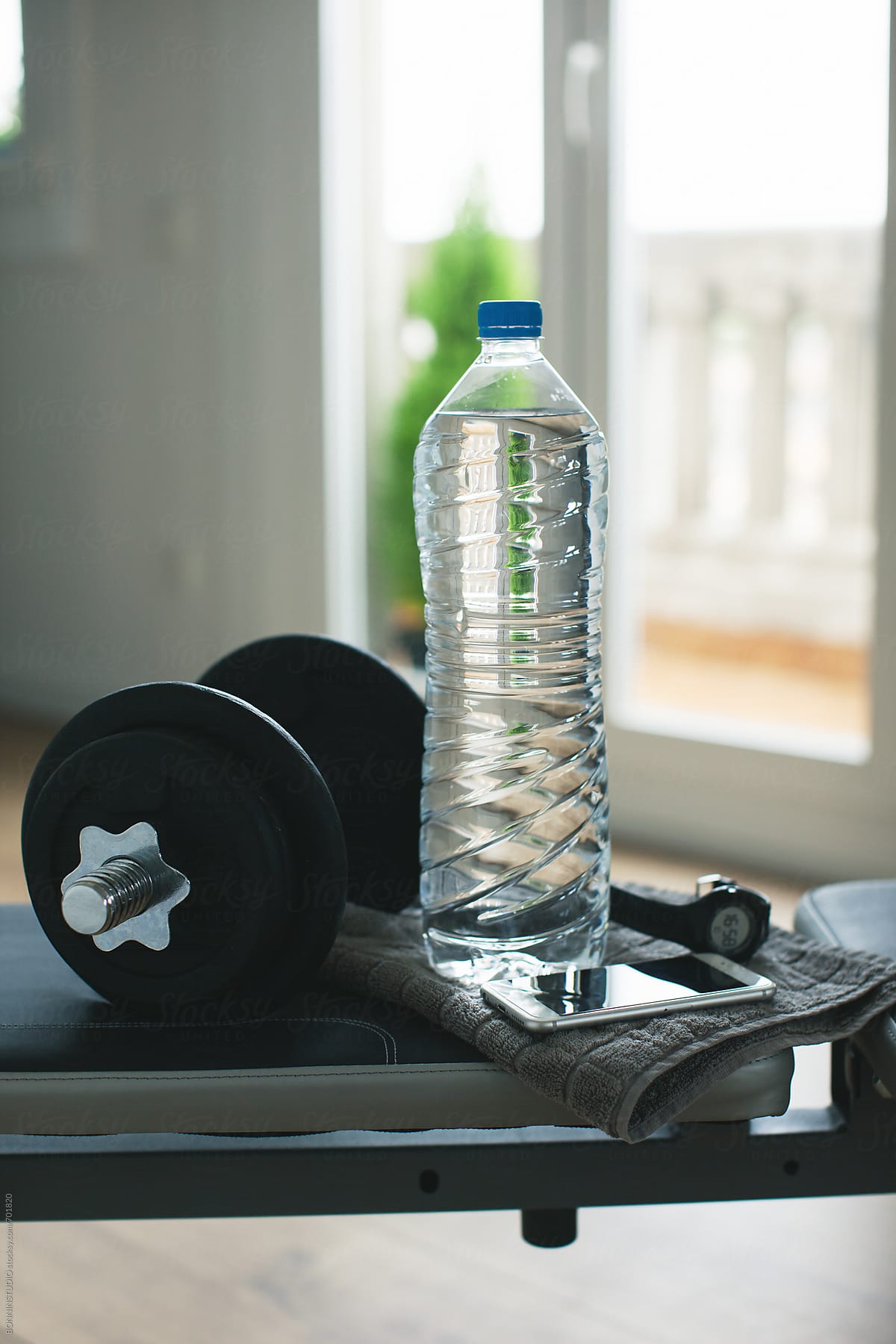 Still life of a weights, a bottle of water, a wristwatch and a phone on a workout bench at home.