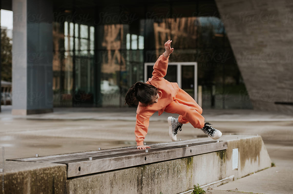 Young girl in runners jumps over concrete bench.