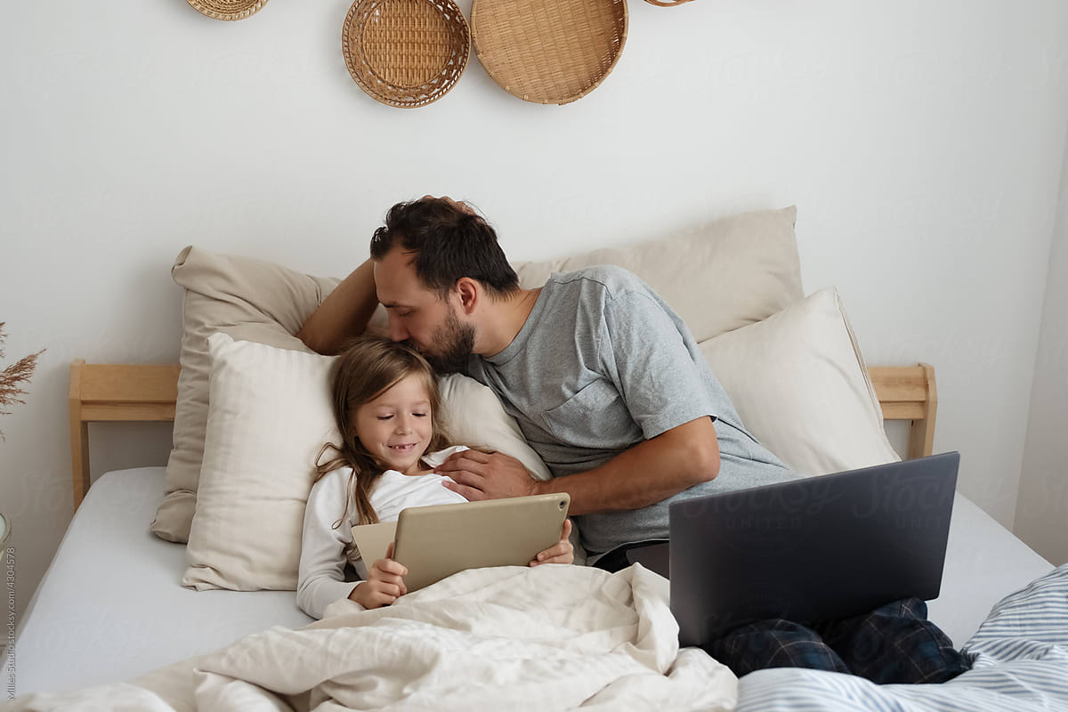 Father kissing daughter with tablet on bed