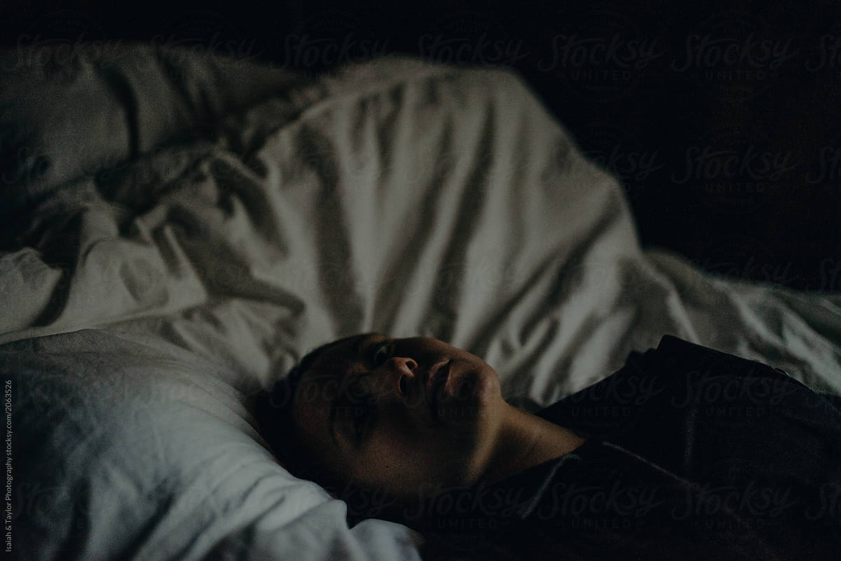 A Dark Dusk Photo Of A Woman Laying On A White Bed With A Shadowed Face Looking At The Camera 