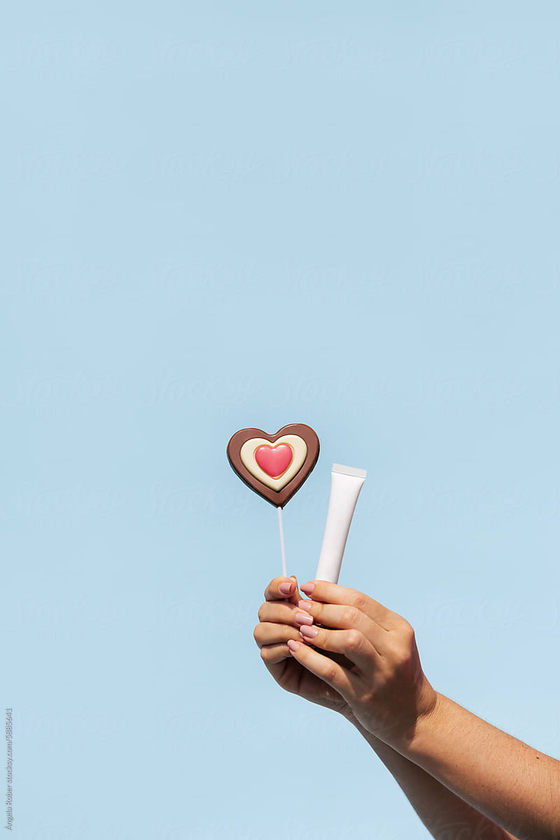 Hands holding a heart-shaped chocolate lollipop and a beauty product