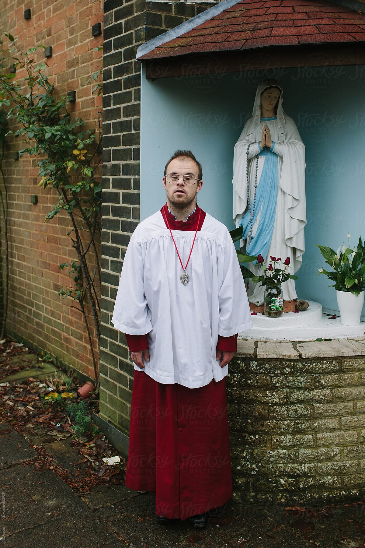 A man with Down\'s Syndrome wearing altar serving robes poses next to a statue of the Virgin Mary.