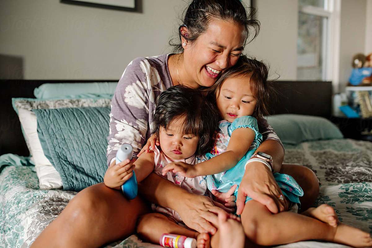 Joyful Asian mom on bed hugging daughters