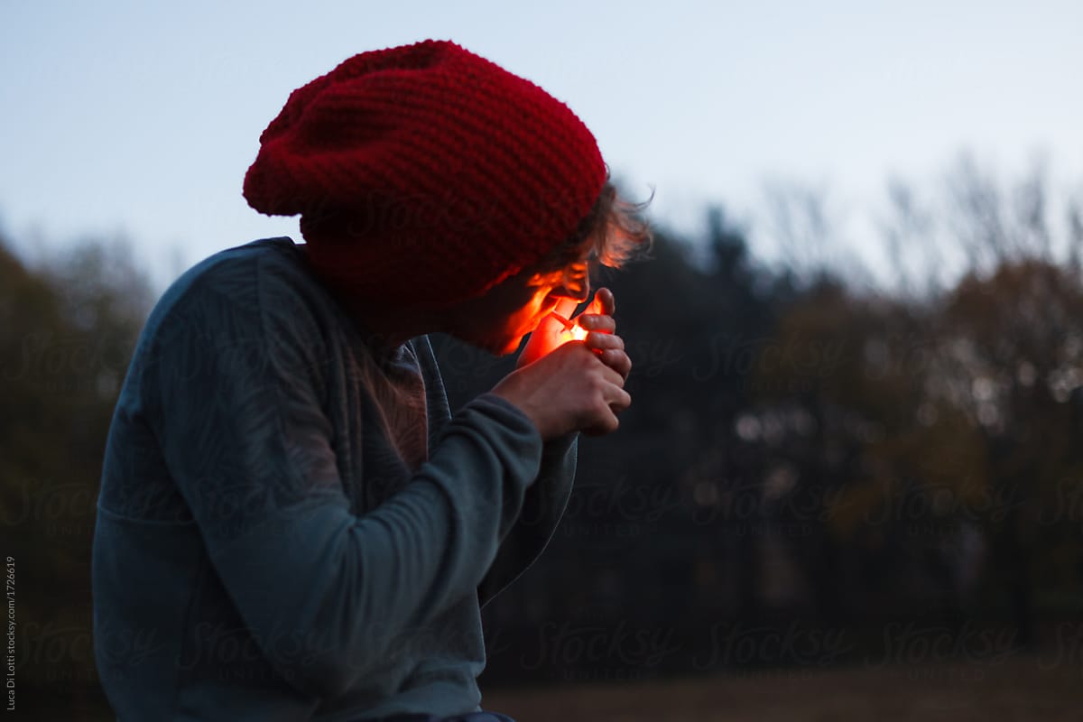 young man wearing a red hat lights a cigarette in a field of a city park