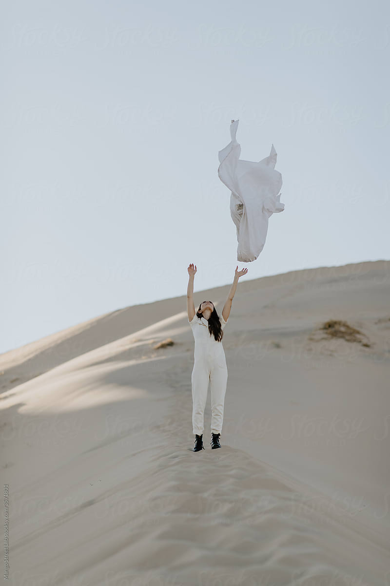Girl in White Jumpsuit and Black Boots with Long Dark Hair Standing on a White Desert Sand Dune Tossing a White Sheet  Against a White Sky