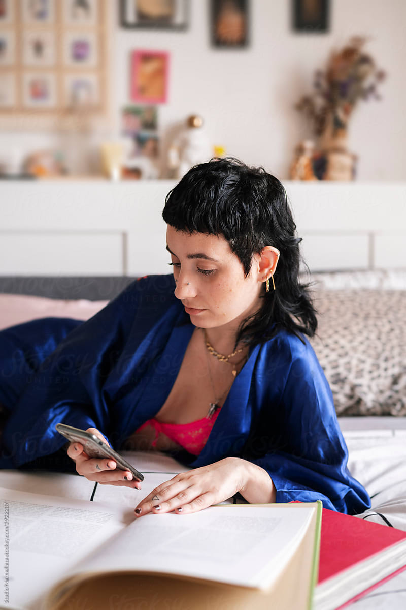 Female student in sleepwear browsing smartphone and reading book