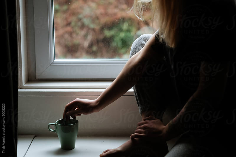 sleepy blonde woman sitting in the window taking a cup of coffee in the morning