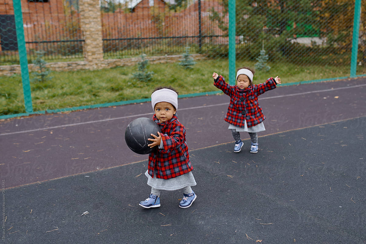 little twin girl in autumn clothes playing with a black basketball on the Playground