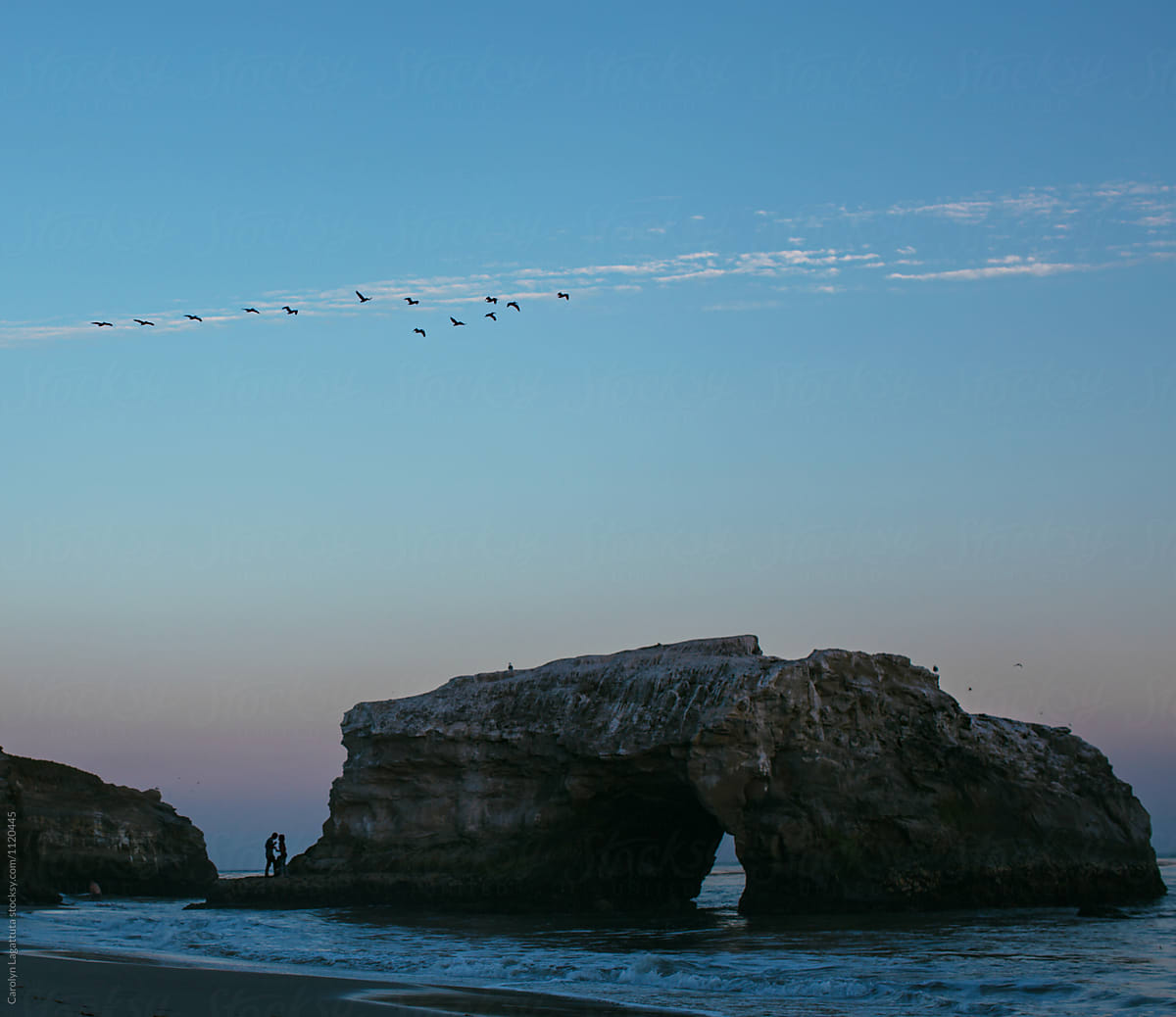 Couple Next To A Natural Bridge Rock In The Ocean At Sunset By