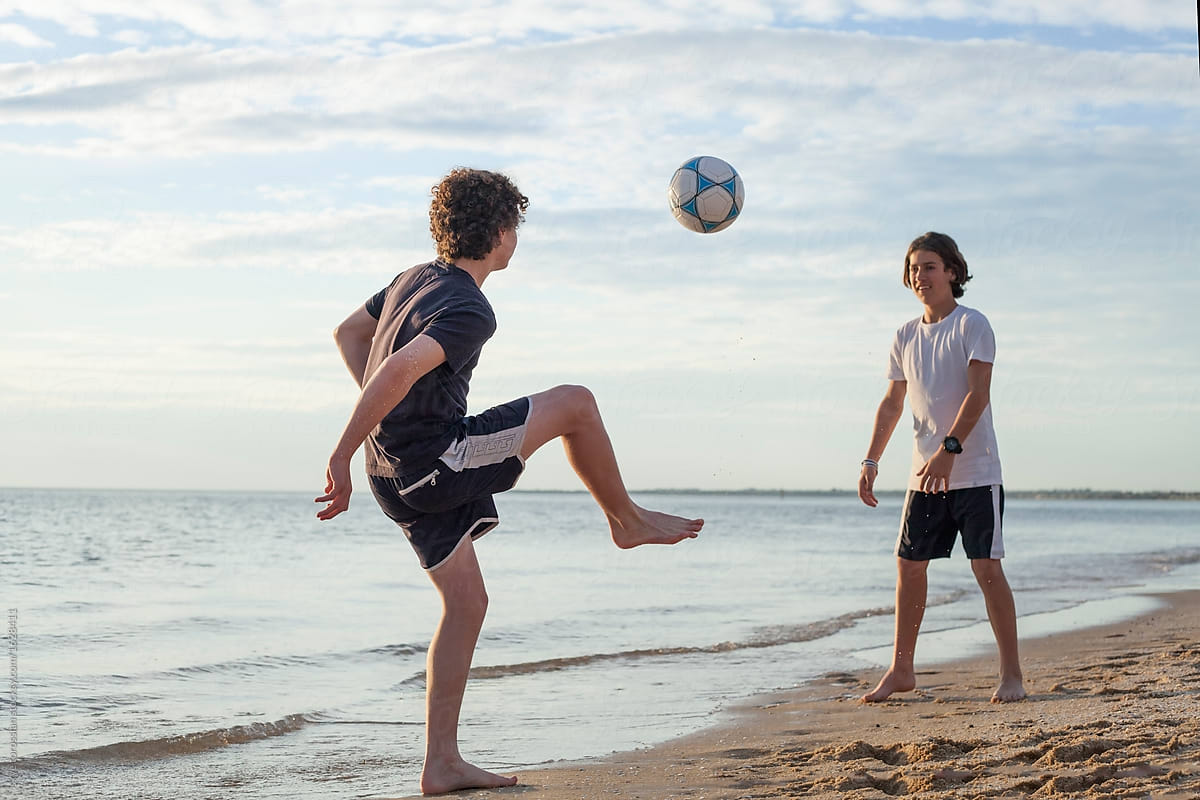 two teen boys juggling a soccer ball on the beach at sunset. 