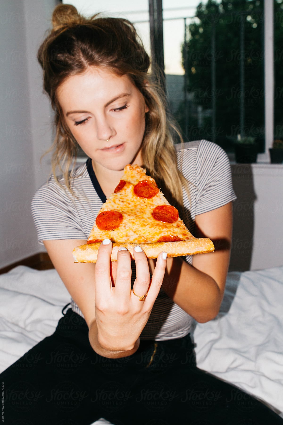 young woman eating slice of pepperoni pizza on bed in apartment